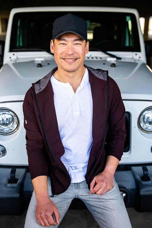 A photo of Aaron smiling and sitting on the front bumper of his Jeep.